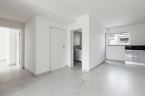Bright empty apartment interior with white walls, marble-effect floors, doorways, windows, and a compact kitchen.
